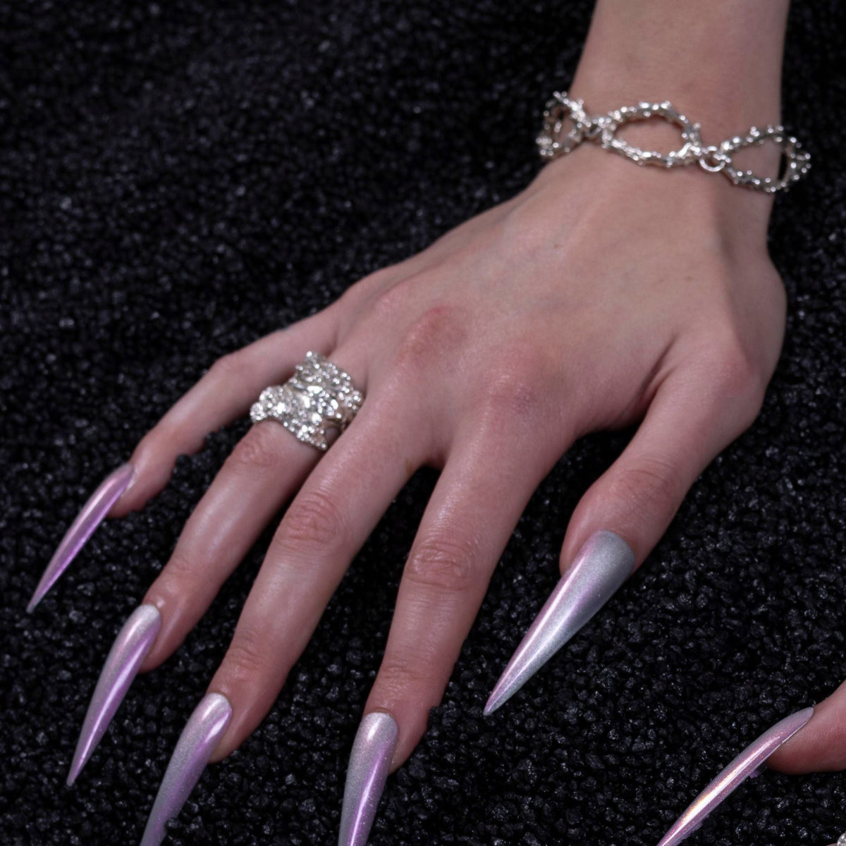 A close-up of a hand with long, sharp stiletto-style nails in a metallic lavender-purple shade, wearing a large rhinestone ring and a silver chain bracelet, set against a dark textured background.